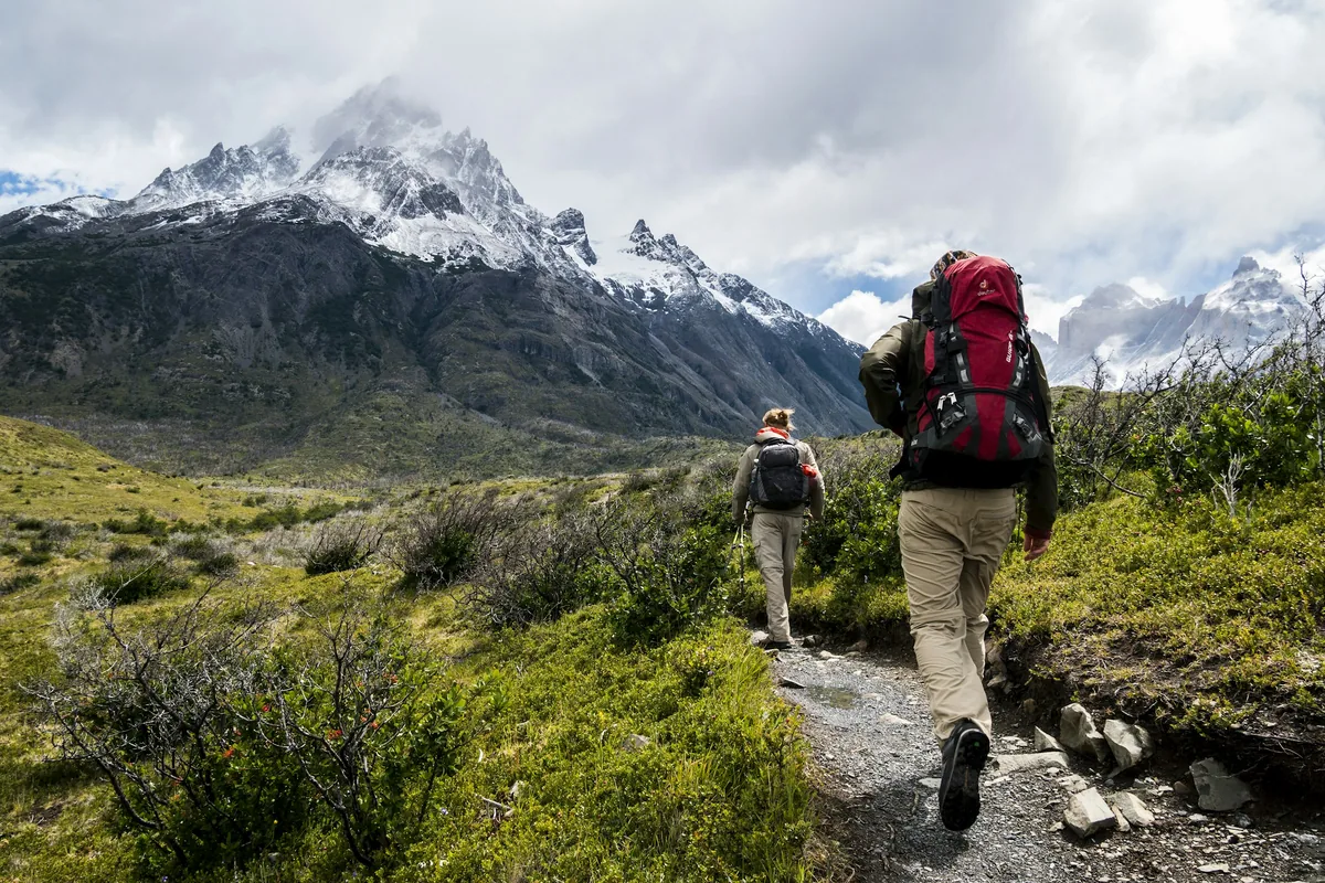 Two hikers with backpacks walk side by side along a trail toward snow-capped mountains in Patagonia.