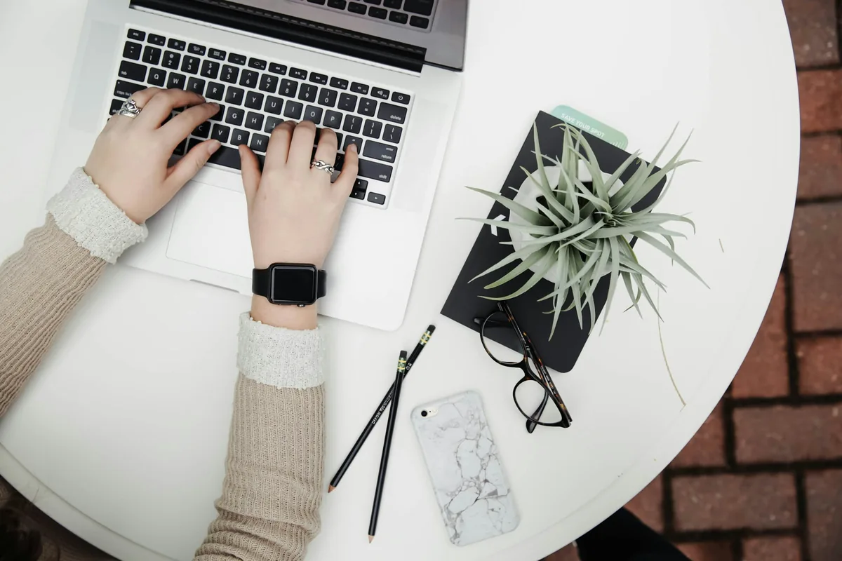 Hands type on a laptop at a white desk beside a potted aloe plant, a phone, and a pair of eyeglasses.