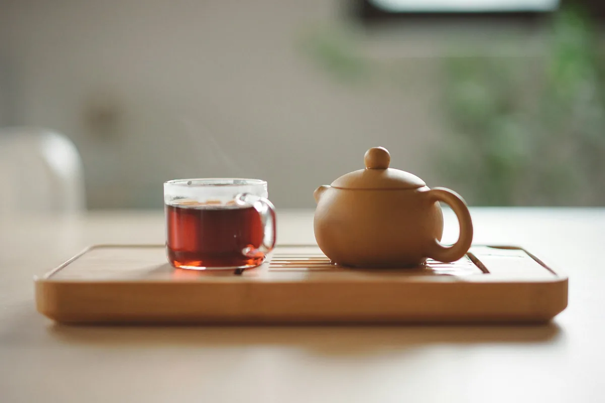 A clear glass cup of tea sits beside a brown ceramic teapot on a table.